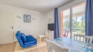Una habitación con sofá azul, una mesita, un televisor sobre un mueble bajo y un balcón con vistas a árboles.