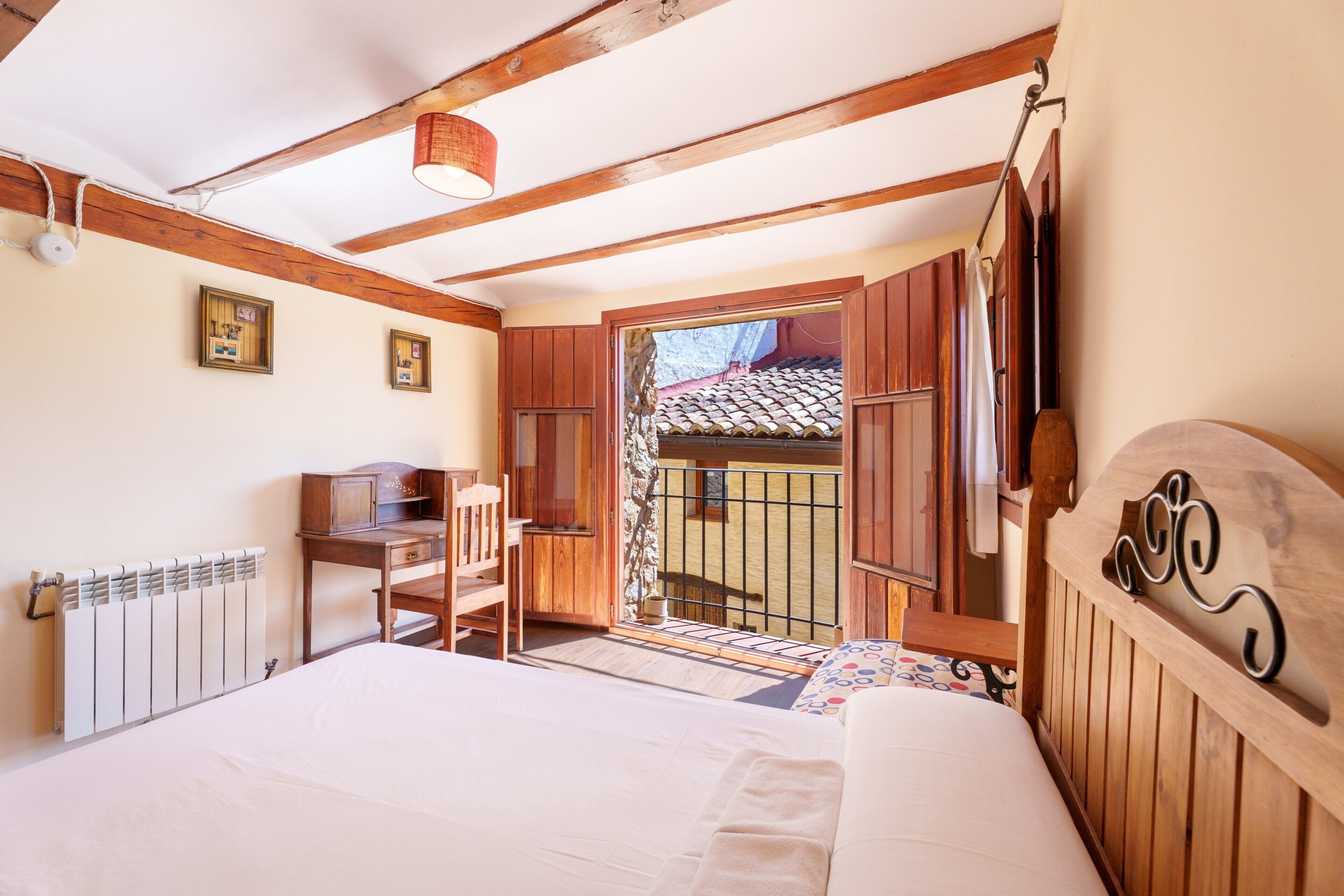 Un dormitorio con cama, escritorio de madera, silla, radiador y balcón con vistas.