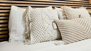 Ein Bett mit weißer Bettwäsche und Makramee-Kissen, ein Holterslatten-Kopfende und ein Wandbehang aus Makramee.
