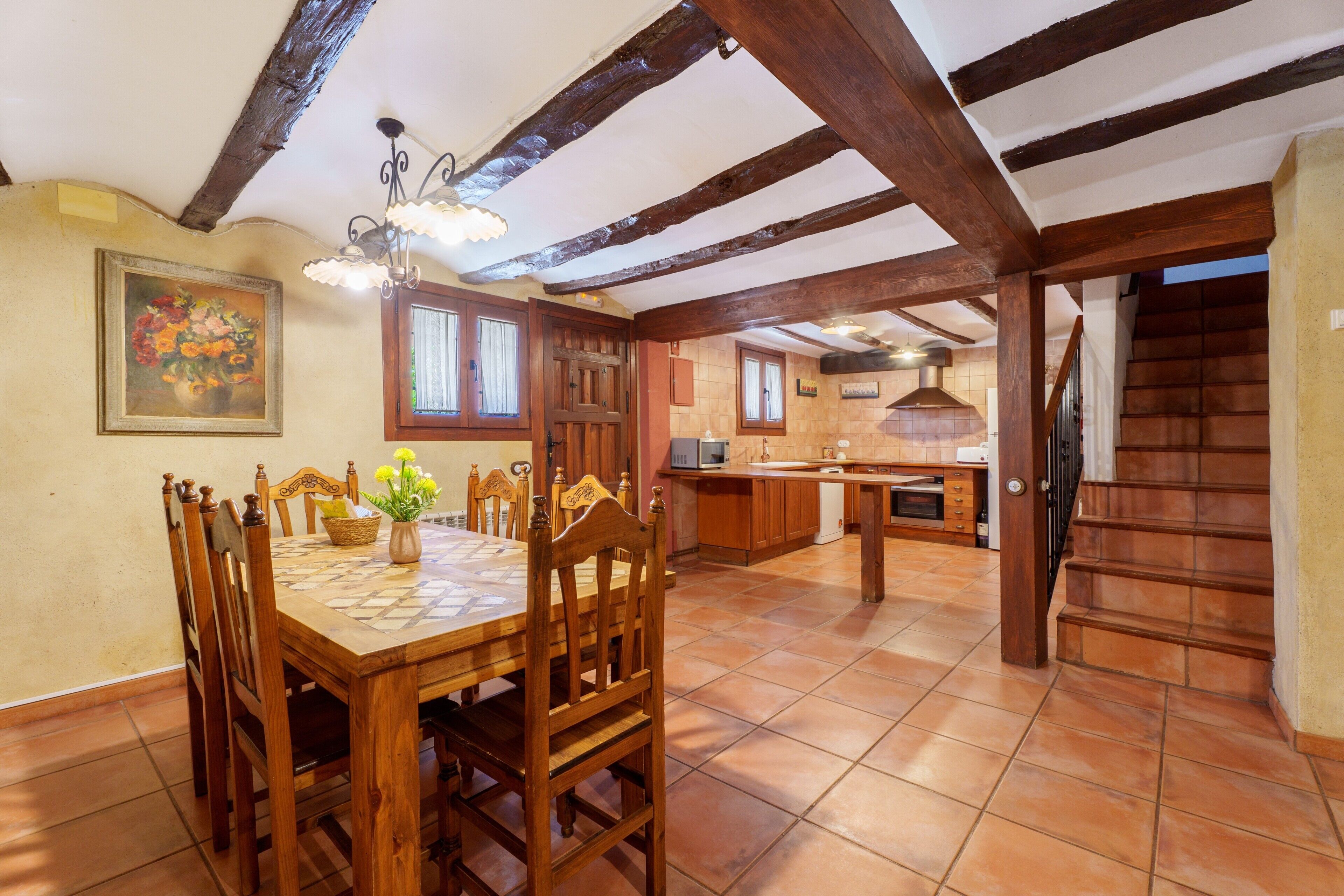 Un coin repas avec une table et des chaises en bois, une cuisine à l’arrière-plan et un escalier menant à un autre niveau.