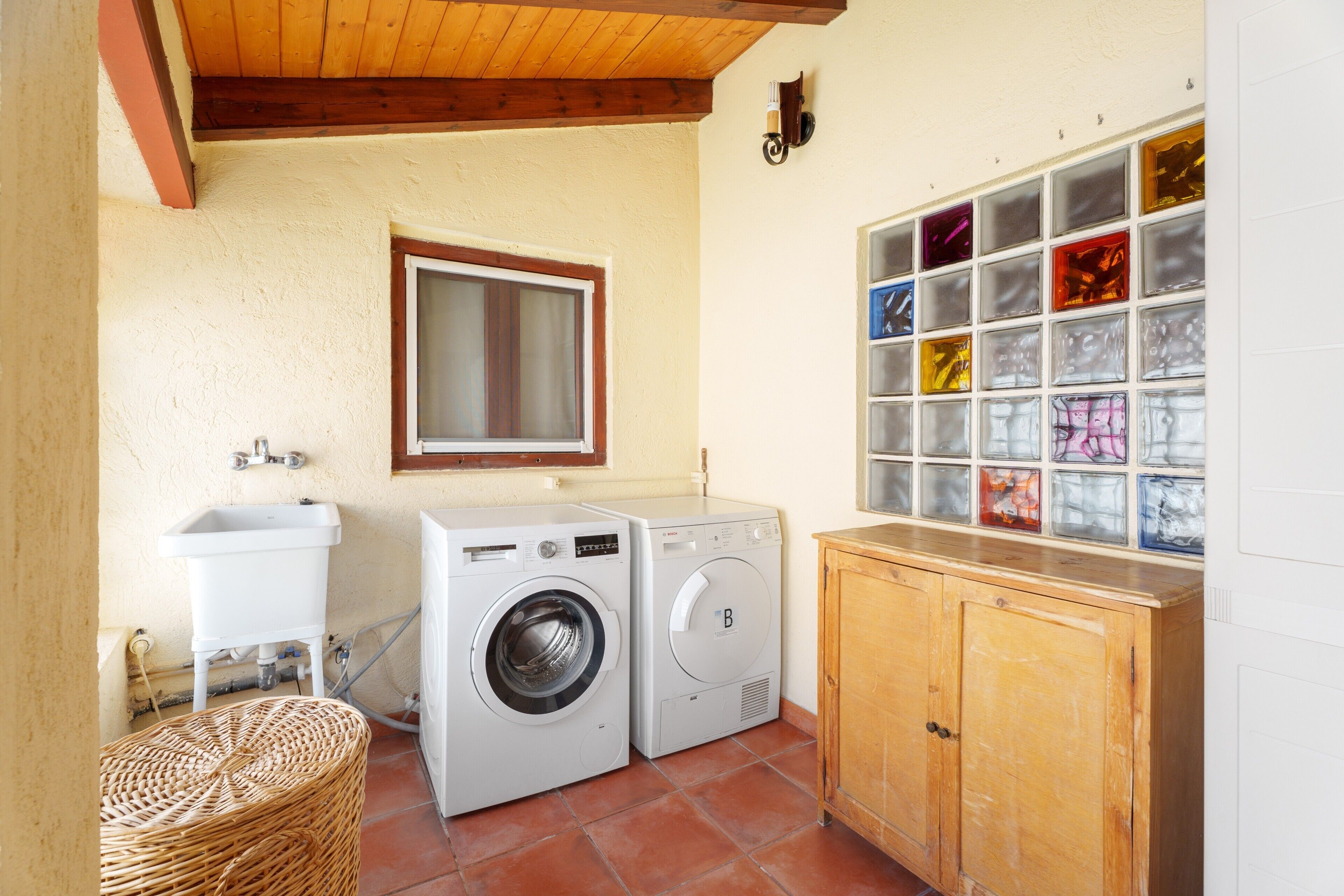 Un cuarto de lavado con lavadora, lavamanos, un gabinete de madera y ventana con vista a bloques de vidrio de colores.