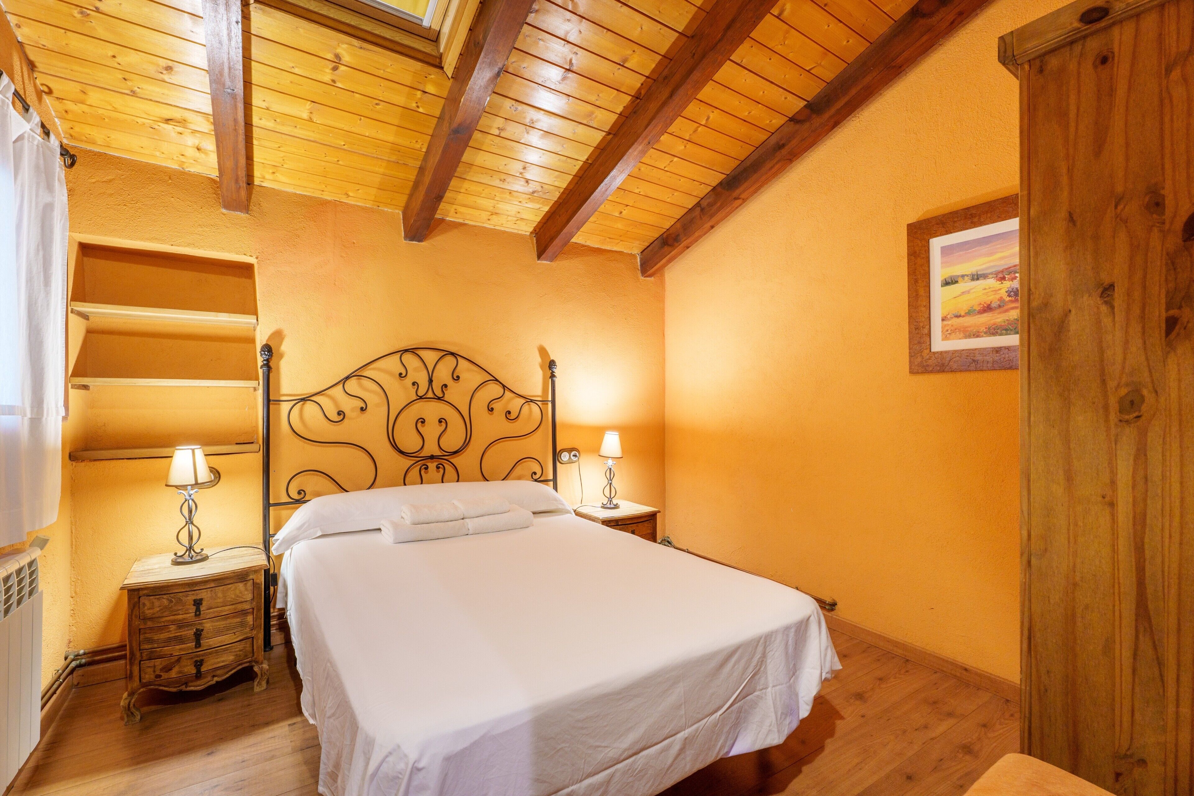 A bedroom with a wooden ceiling, a bed with white linens, two bedside tables with lamps, and a framed picture on the wall.