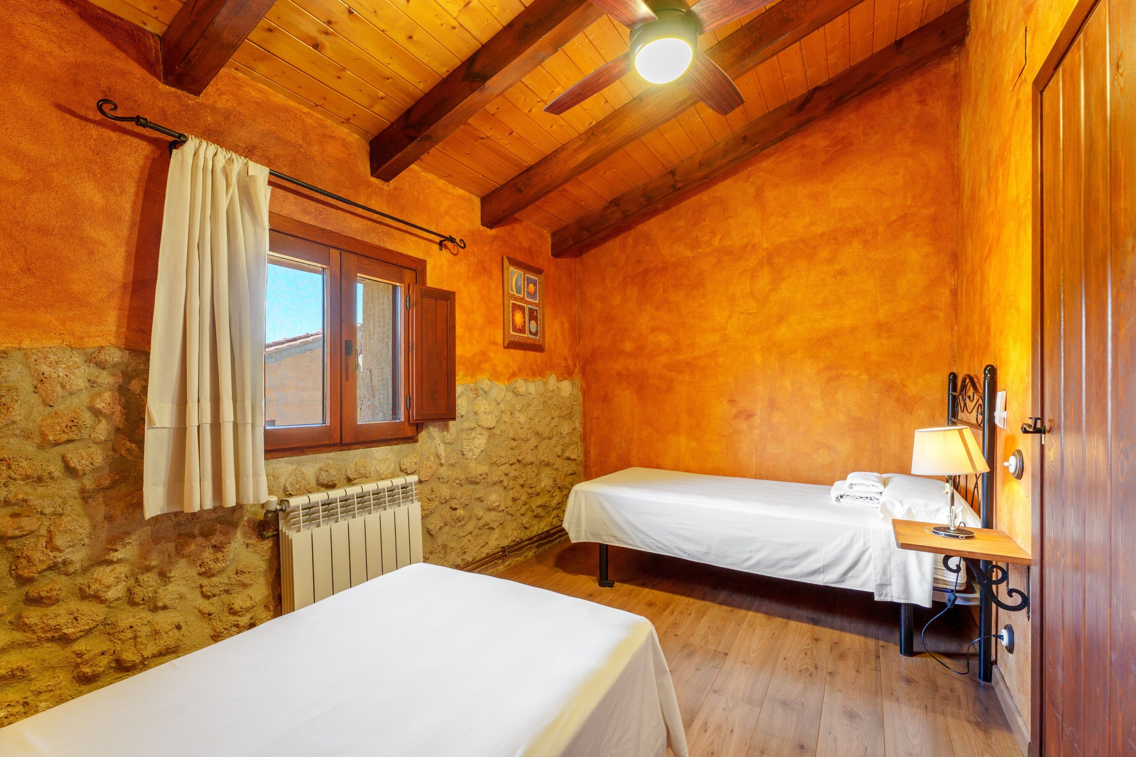 Ein Zimmer mit zwei Betten, einem Vorhangfenster, einem Heizkörper, einer Holzbalkendecke und einer Wandlampe.