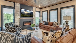 A modern living room with a fireplace, leather sofas, patterned armchairs, and large windows offering a view of the outdoors. - Special Offer•6BD•Pool•Theater•Games•Views (Sevierville)