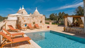 Un área junto a la piscina con sillones de descanso, un edificio de piedra con techos cónicos y un cielo azul despejado.