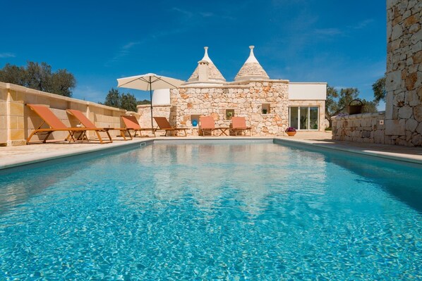 A swimming pool with lounge chairs, a stone building with conical roofs, and a clear blue sky. - Holiday Home 'Trullo Amami With Private Pool' with Private Pool, Wi-Fi and Air Conditioning (san michele salentino)