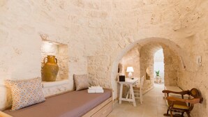 Una habitación con paredes de piedra, una cama, un banco de madera, una mesa con una lámpara y un florero en una repisa.