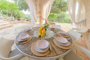 A round dining table with white chairs, set with plates, cutlery, and a centerpiece of lemons and bananas.