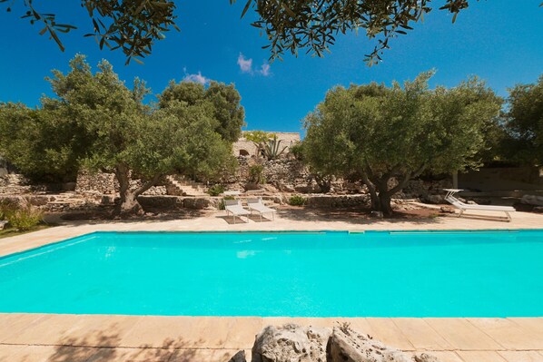 A swimming pool with lounge chairs, surrounded by olive trees and stone structures.