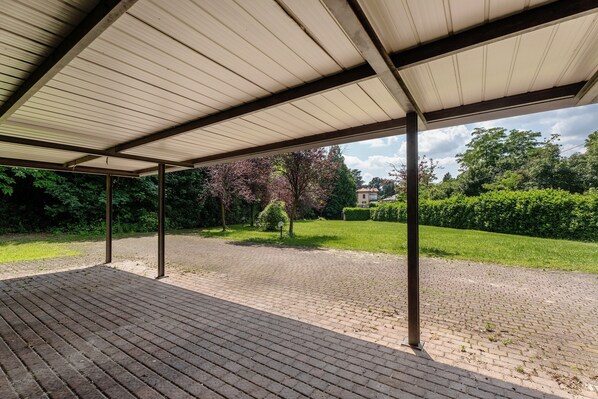 A covered outdoor area with a brick floor, metal beams, and a view of a green landscape with trees and a building in the background.