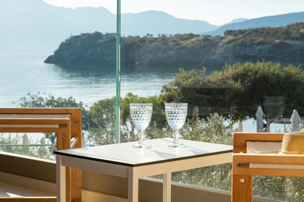 A table with two clear glass cups, a wooden chair, and a view of the sea and mountains.