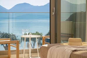 A balcony with a glass railing overlooking a body of water and mountains. There is a bed with a beige blanket and a straw hat on it.
