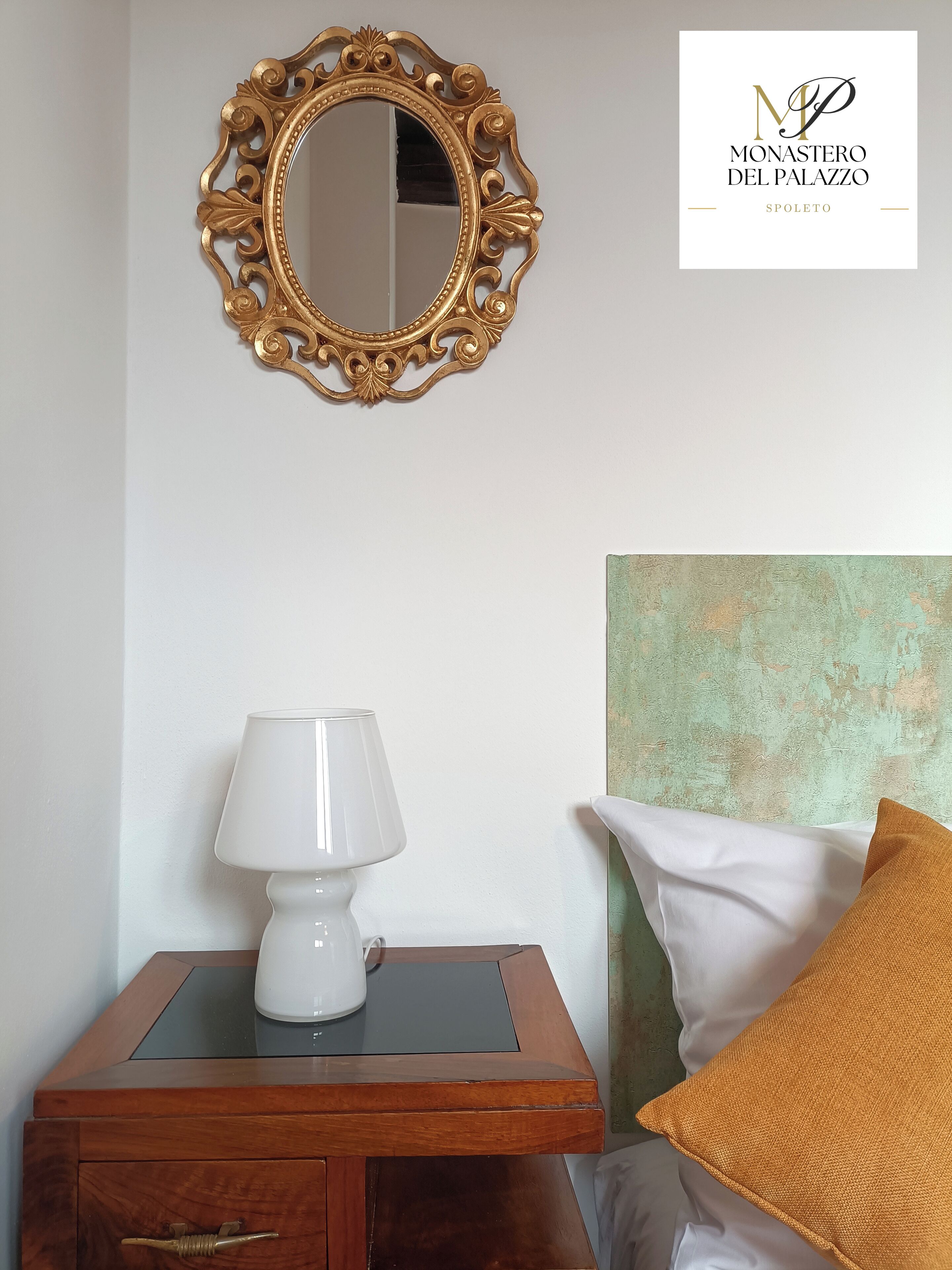 Une chambre avec une table de chevet en bois, une lampe blanche, un miroir à cadre doré, un oreiller blanc et un mur à texture.