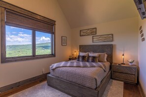 A bedroom with a large window, a wooden headboard, a bedside table with a lamp, and a view of a green landscape. - Bella Vista at Eagles Nest (Banner Elk)