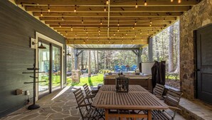 Un patio al aire libre con suelo de piedra, mesa y sillas de madera, y luces colgantes.