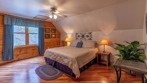 Un dormitorio con piso de madera, una cama con cabecera, dos mesitas de noche con lámparas, un ventilador de techo y una ventana con cortinas.