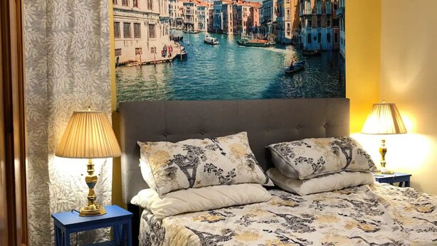 A bedroom with a bed, two bedside lamps, a blue nightstand, a patterned bedspread, and a large cityscape painting on the wall. - Cozy Mediterranean Escape  (Schenectady)