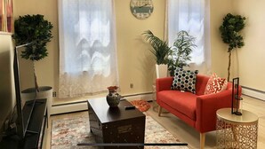 A living room with a red sofa, two potted plants, a clock on the wall, and a large window with curtains. - Cozy Mediterranean Escape  (Schenectady)
