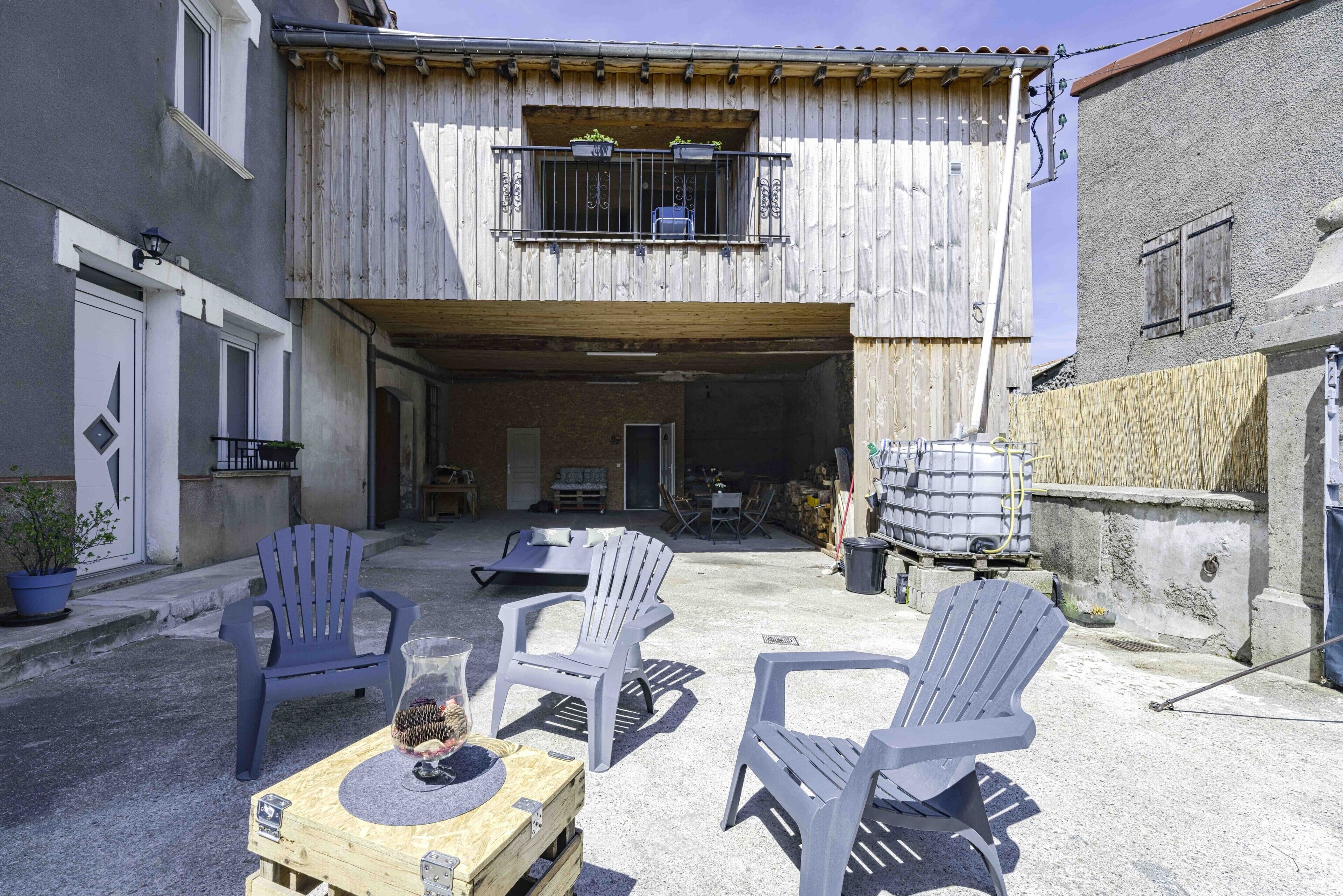 Um edifício com varanda de madeira, área de estar externa com cadeiras e mesa, e entrada para garagem.
