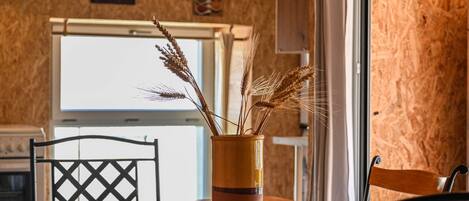 Una mesa de madera con un florero de espigas de trigo, una silla negra y una ventana que da a una cocina.