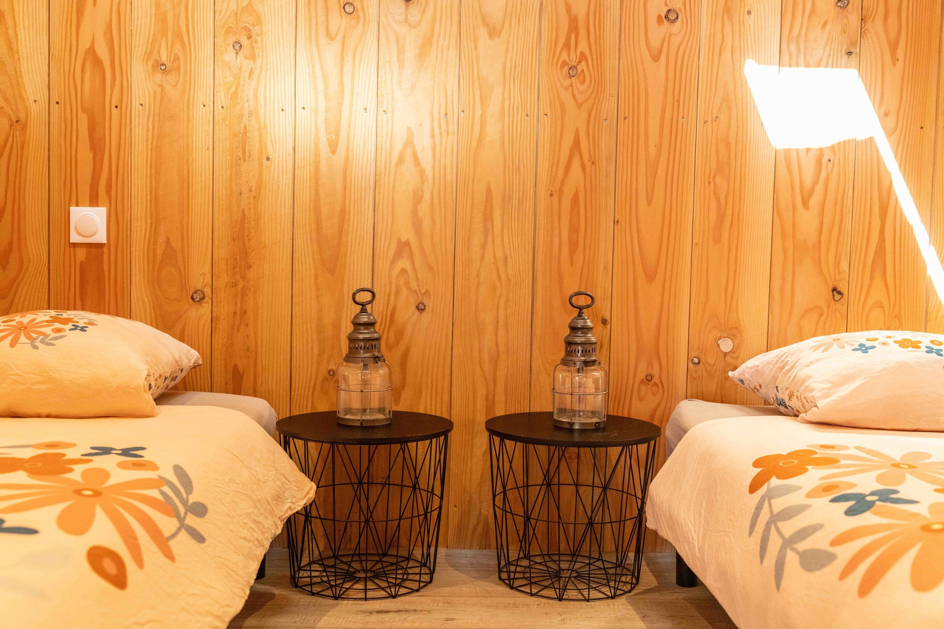 Een kamer met houten lambrisering, met twee bedden, elk voorzien van een bloemenprint beddeken, twee kleine ronde tafels met een metalen draadframe, en twee glazen potten op de tafels.