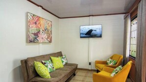 Ein Wohnzimmer mit einem braunen Sofa, zwei gelben Sesseln, einem an der Wand montierten Fernseher und einem floralen Gemälde über dem Sofa.