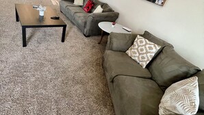Una sala de estar con un sofá gris, una mesa de centro y una mesita blanca. Se ve una cocina al fondo con una silla roja.