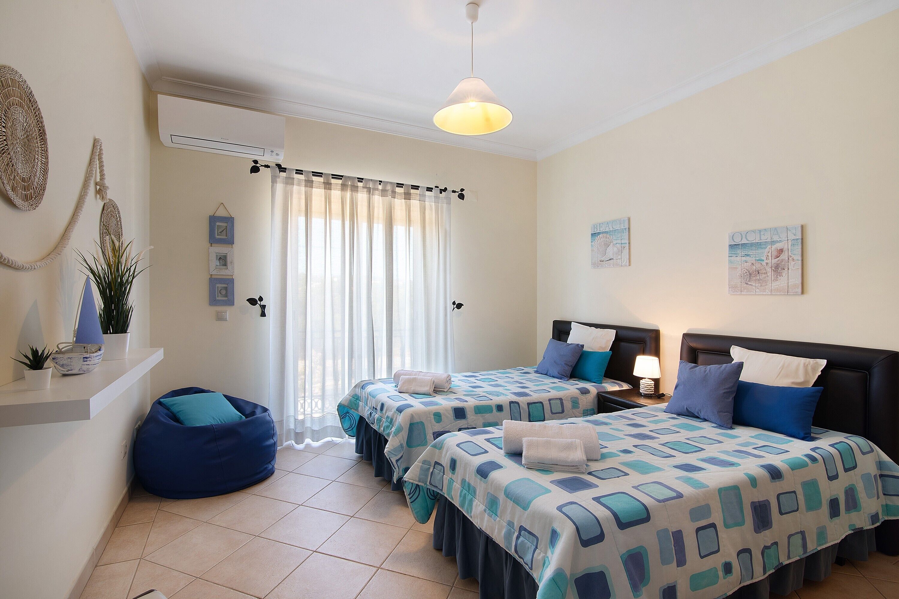 A hotel room with two beds, a ceiling fan, a window with curtains, a bedside table with a lamp, and wall art.