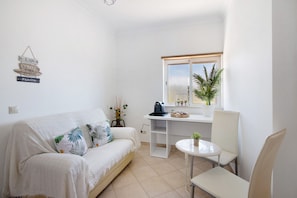 A small, clean room with a white sofa, a small table, a chair, a window with a view, and a wall-mounted sign. - Villa 'J R Dream House' with Private Pool, Wi-Fi and Air Conditioning (Quarteira)