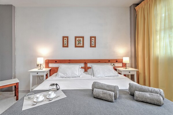 A hotel room with a bed, two bedside tables, a lamp, a bedside table with a cup and saucer, a tray with a cup, a glass, and a spoon.