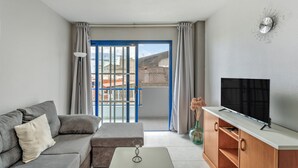 Un salón moderno con un sofá, una mesa de centro, un televisor en un mueble de madera y un balcón con puertas azules.