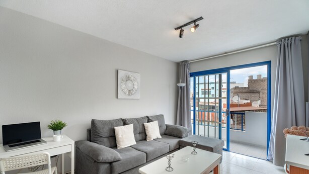 Una sala de estar moderna con un sofá gris, una mesa de centro blanca, una computadora portátil sobre un escritorio y vistas a través de una ventana con marco azul.