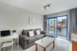 A modern living room with a grey sofa, a white coffee table, a laptop on a desk, and a view through a blue-framed window.