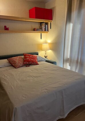 A neatly made bed with a white bedspread and two red pillows, a bedside table with a lamp, a red cabinet above the bed, and a window with curtains.