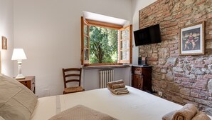 A bedroom with a large bed, a window with an open shutter, a flat-screen TV mounted on a brick wall, and a radiator below the window. - Country House 'La Pineta Farm House' with Private Pool and Wi-Fi (Varna)