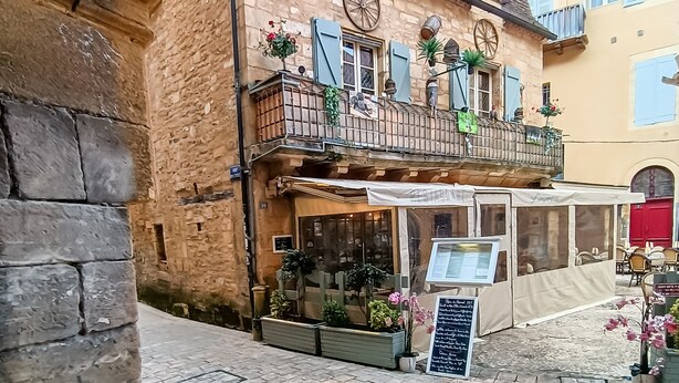 A stone building with a balcony, a cafe with outdoor seating, and a signboard.
