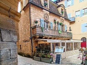 A stone building with a balcony, a cafe with outdoor seating, and a signboard.