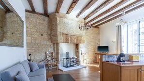 A rustic interior with exposed stone walls, wooden beams, a fireplace, a sofa, a coffee table, and a kitchen area.