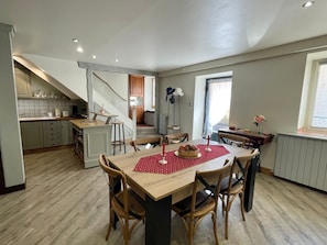 A dining area with a wooden table and chairs, a kitchen in the background, and a staircase leading to another level.