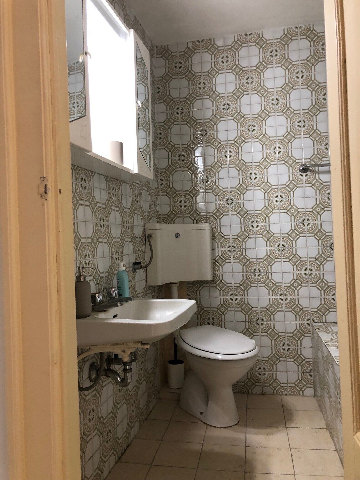 A small bathroom with a toilet, sink, and mirror. The walls are tiled with a geometric pattern.