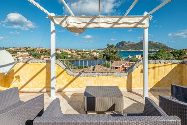 Terraza en la azotea con mobiliario de mimbre, una pérgola y vistas a un pueblo costero y al mar.