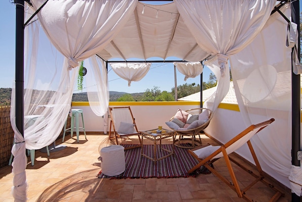 A covered outdoor seating area with wicker chairs and a table, a white canopy, and a view of the landscape.