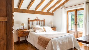A bedroom with a wooden ceiling, a large window with a view of greenery, a bed with a white bedspread, and a nightstand with a lamp.