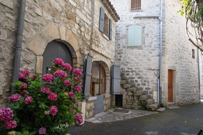 Um edifício de pedra com janelas em arco e uma porta, uma pequena escadaria e um arbusto de flores em primeiro plano.
