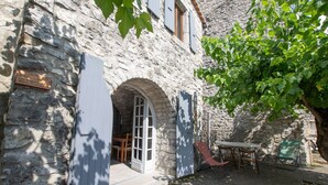 A stone building with an arched doorway, a table with chairs, and a small garden area. - Holiday Home 'Gite De L'écurie' with Air Conditioning (Berrias-et-Casteljau)