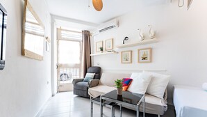 Ett modernt vardagsrum med en soffa, en fåtölj, ett soffbord och en säng. Det finns vägghängda hyllor med dekorativa föremål och ett fönster med gardiner.