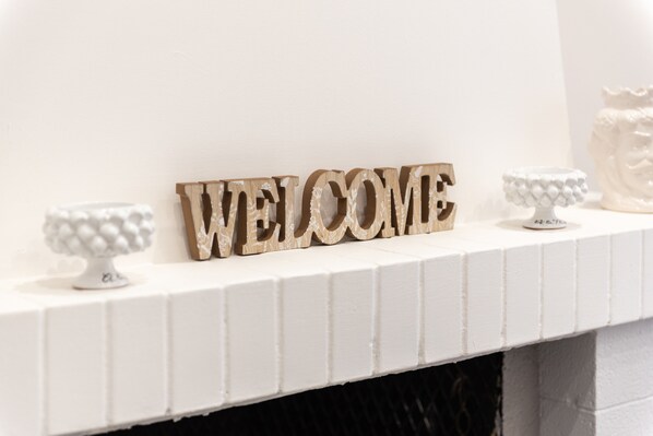 Ein weißer Kaminvorsatz mit einem Holzschild mit der Aufschrift 'WELCOME' und zwei dekorativen Kerzenhaltern.