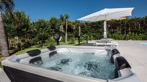 Un bain à remous doté d’un parasol blanc, entouré d’une végétation luxuriante et d’une piscine.