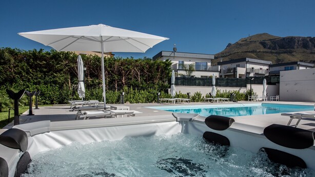 Un espace avec un jacuzzi, des chaises longues et des parasols, une piscine, et des bâtiments modernes en arrière-plan.