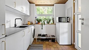 Une cuisine compacte avec des meubles blancs, un évier, un four à micro-ondes et un four encastré. Une fenêtre est agrémentée de plantes posées sur le rebord, et un porte-chaussures se trouve en dessous.